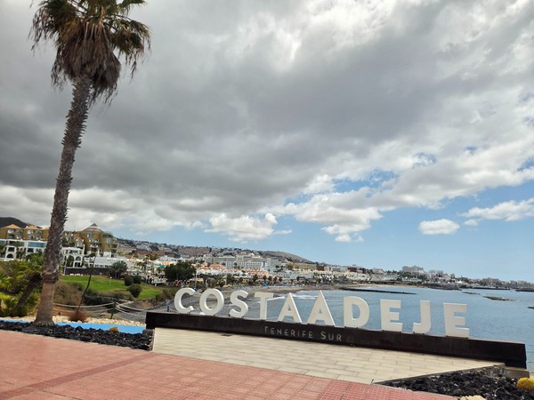 Letrero de Costa Adeje Tenerife Sur junto al mar, con la costa urbana y hoteles al fondo bajo un cielo nublado.