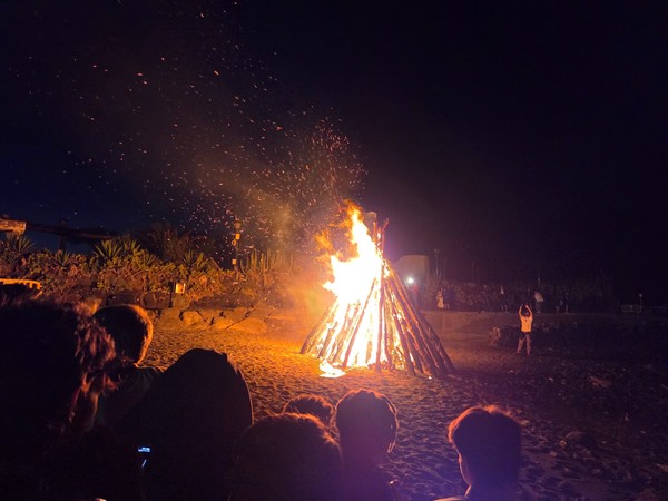Hoguera de la Noche de San Juan ardiendo en la playa, con chispas en el aire y personas observando alrededor.