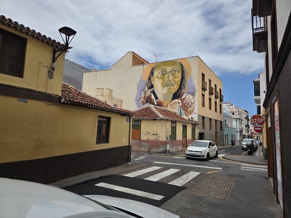 Mural de retrato femenino en una calle de Puerto de la Cruz, rodeado de edificios tradicionales y coches aparcados.