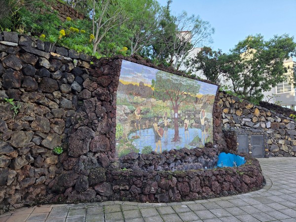 Mosaico cerámico en un muro de piedra volcánica en un parque de Puerto de la Cruz, rodeado de vegetación.