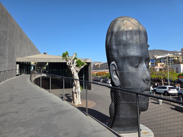 Escultura de una gran cabeza junto al edificio de la biblioteca en Santa Cruz, con una pasarela curva y cielo despejado.